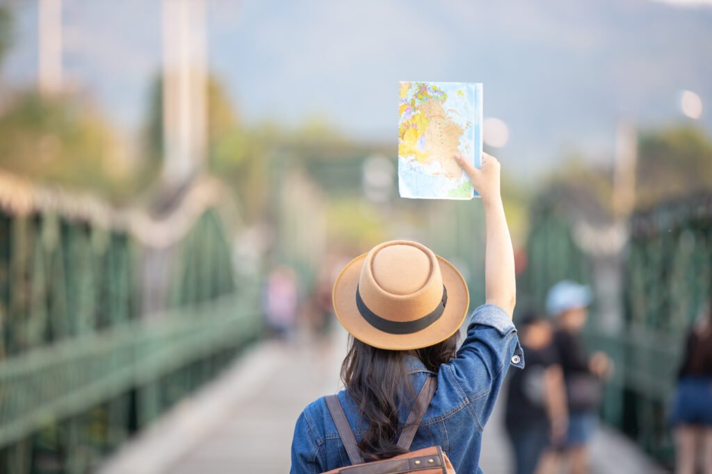 Uma mulher branca de cabelos pretos aparece de costas em uma ponte. Ela veste camisa jeans, uma mochila e um chapéu. Com o braço direito levantado, ela segura um mapa, que representa o uso do inglês para viagem durante visitas a pontos turísticos no exterior.