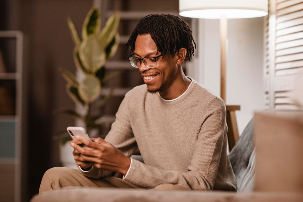 Um homem negro, usando roupas em tons de marrom e óculos, mexendo no celular.