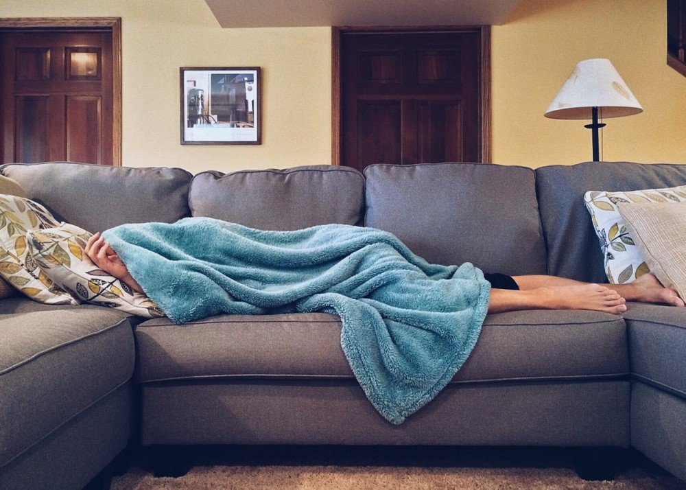Uma pessoa branca deitada em um sofá cinza e coberta com uma manta.