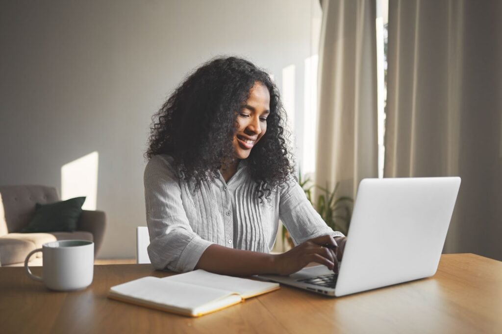 Mulher negra vestindo uma blusa de manga longa cinza e digitando em um laptop prata.