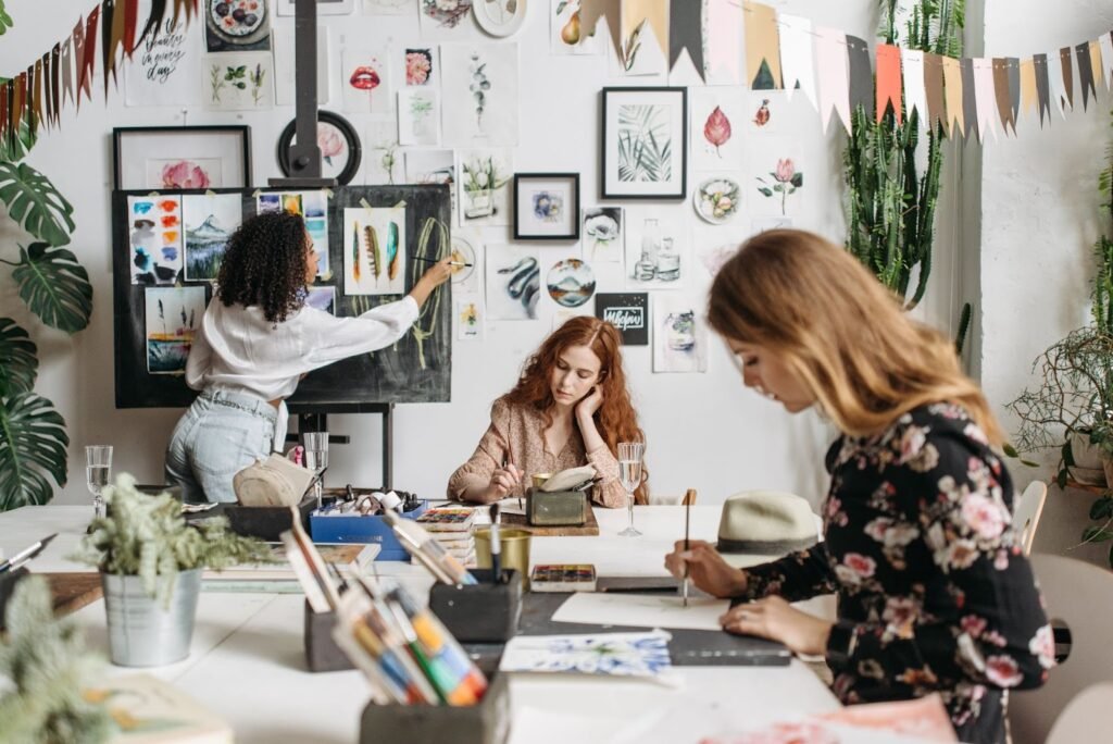 Um escritório com imagens espalhadas na parede e na mesa de trabalho, com duas mulheres sentadas enquanto trabalham concentradas e uma mulher de pé organizando um quadro.