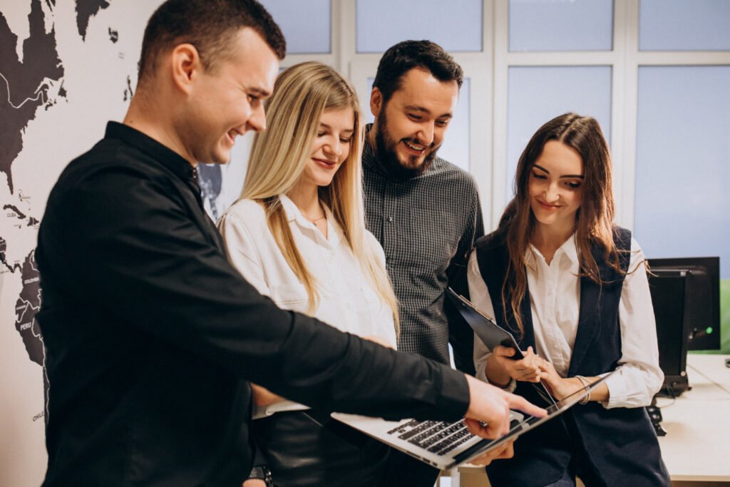 Um grupo formado por quatro pessoas brancas em um ambiente de escritório, observando a tela de um notebook, sorridentes.