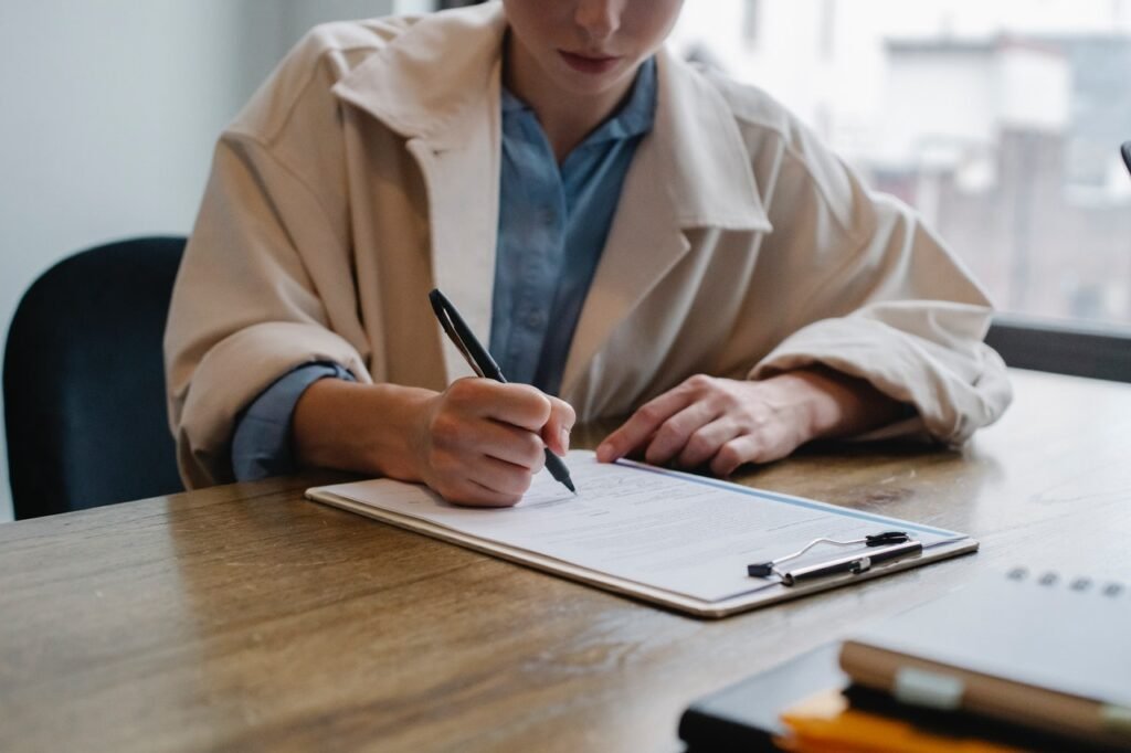 Uma mulher branca está sentada em um escritório. Ela tem uma caneta na mão direita e faz anotações em uma prancheta.