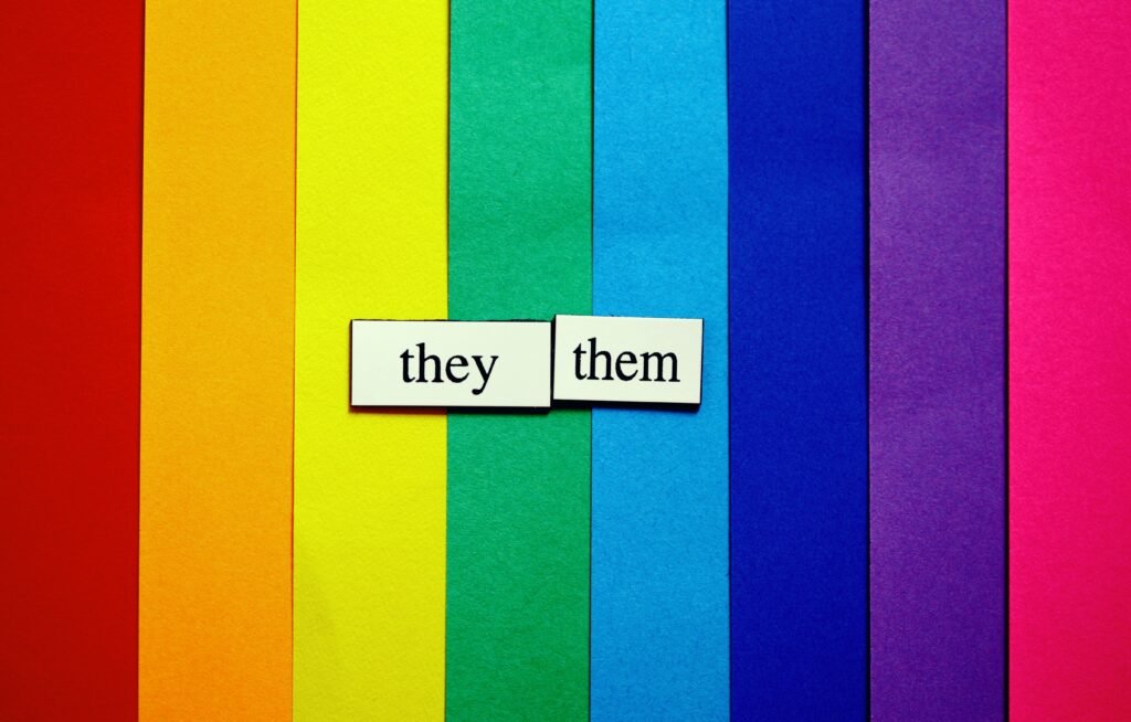 Folhas de papel coloridas formam uma bandeira do arco-íris com duas placas brancas escritas “they” e “them”.