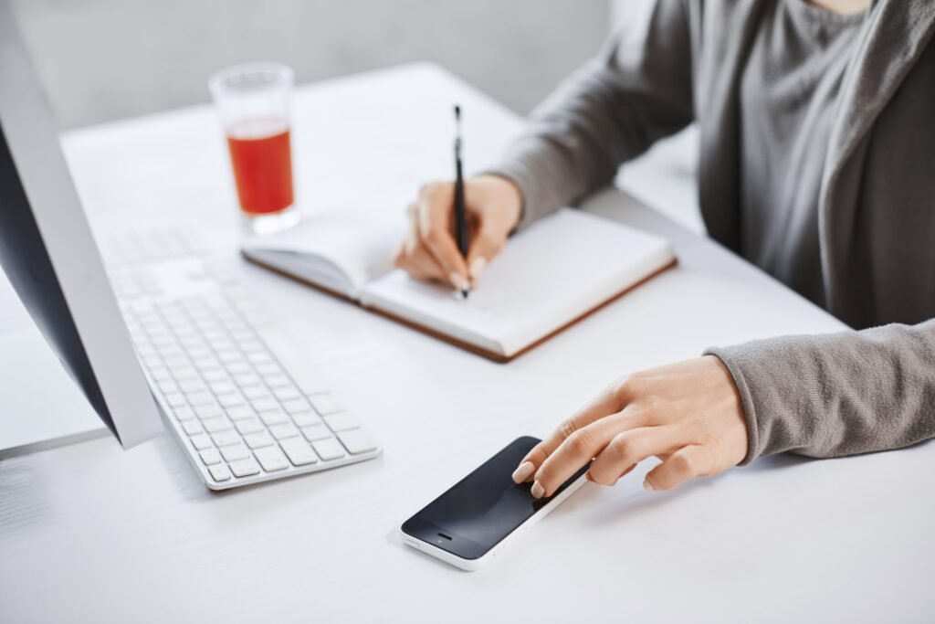 Uma mulher, sentada em frente ao computador, escreve em um caderno apoiado à mesa. Na mesa também estão um celular e um copo de suco.