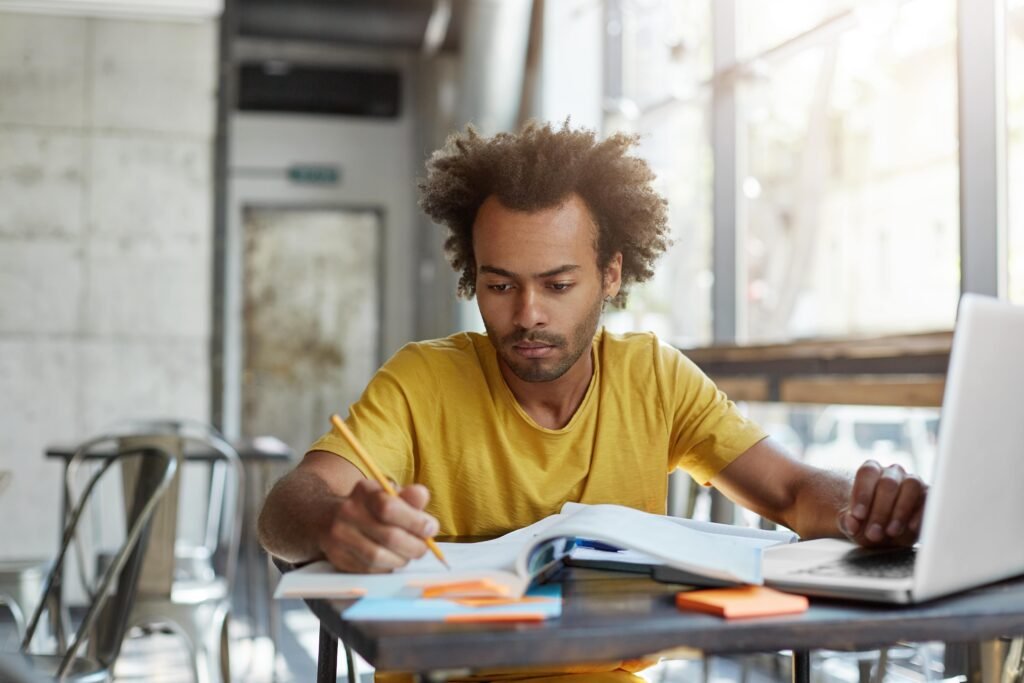 Homem negro usando camiseta amarela sentado em frente a uma mesa. Ele segura um lápis sobre um livro e apoia uma das mãos em um laptop prata.