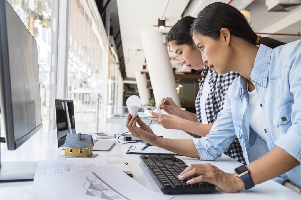 Duas mulheres trabalhando em uma lâmpada e um conector, em frente a um monitor e um laptop sobre uma mesa com alguns papéis e anotações.