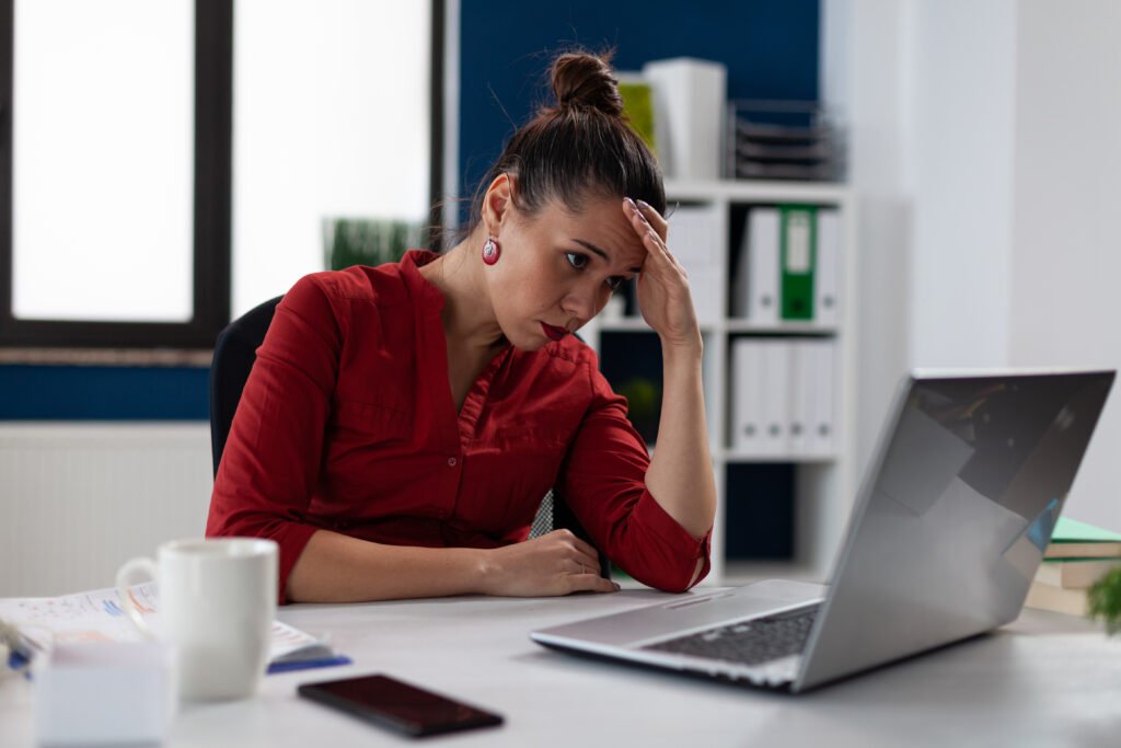 Mulher usando camisa, batom e brincos vermelhos, sentada em frente a um laptop com uma mão na cabeça e expressão chateada.